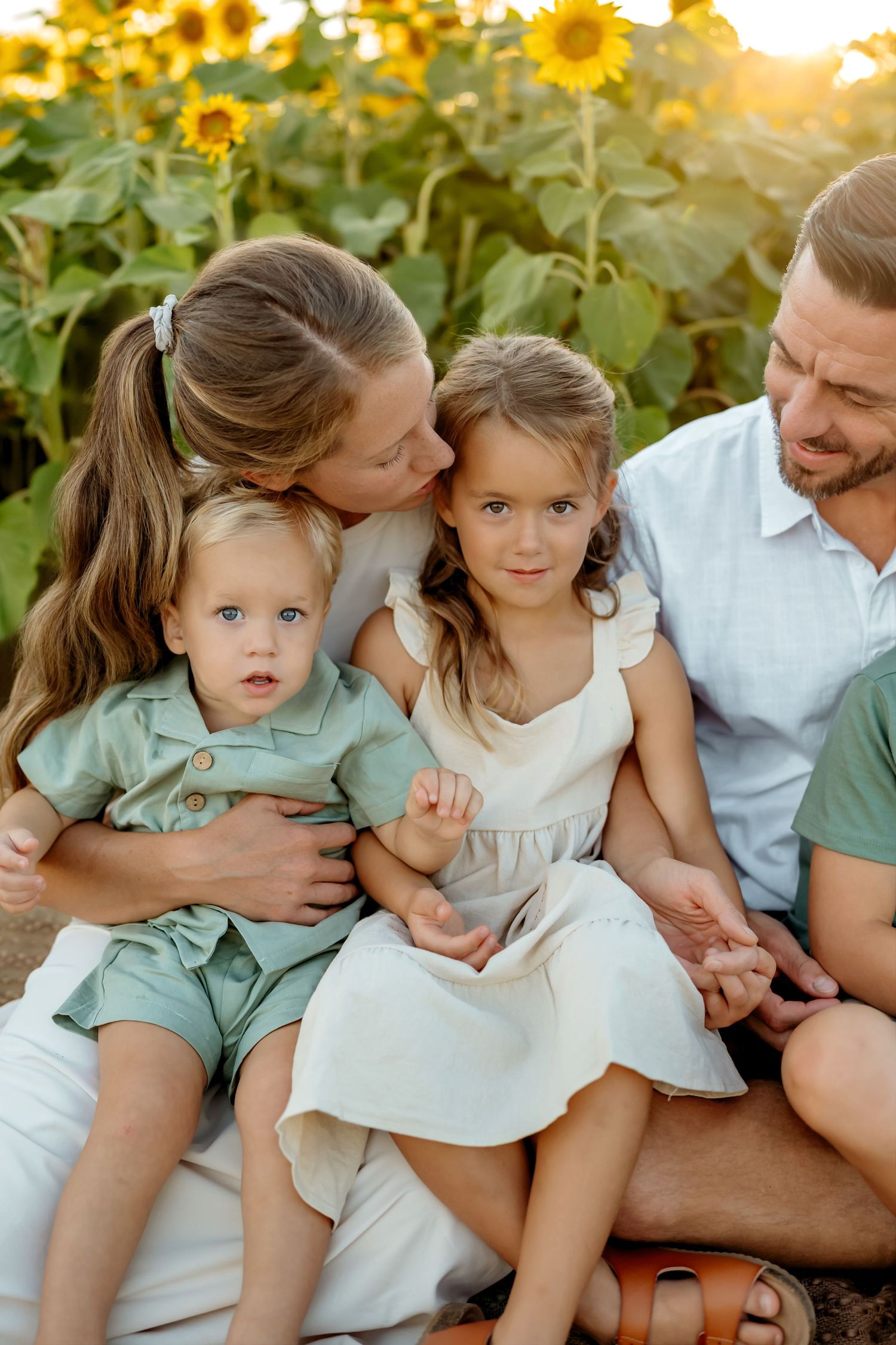 Family lounging together in coordinated mint green and white outfits by blooming sunflowers during golden hour.