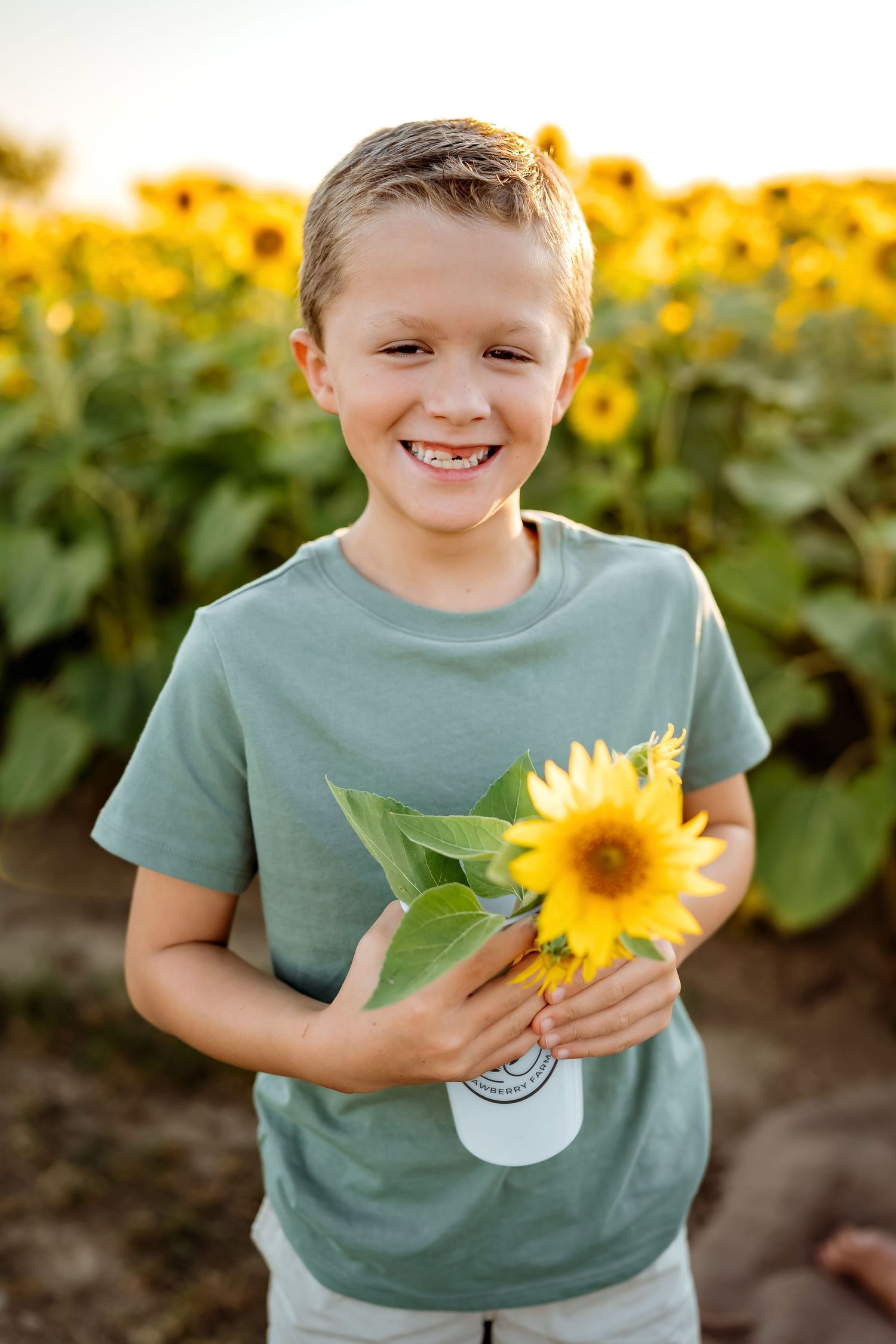 Young child in sage green shirt holding sunflower while smiling in garden.