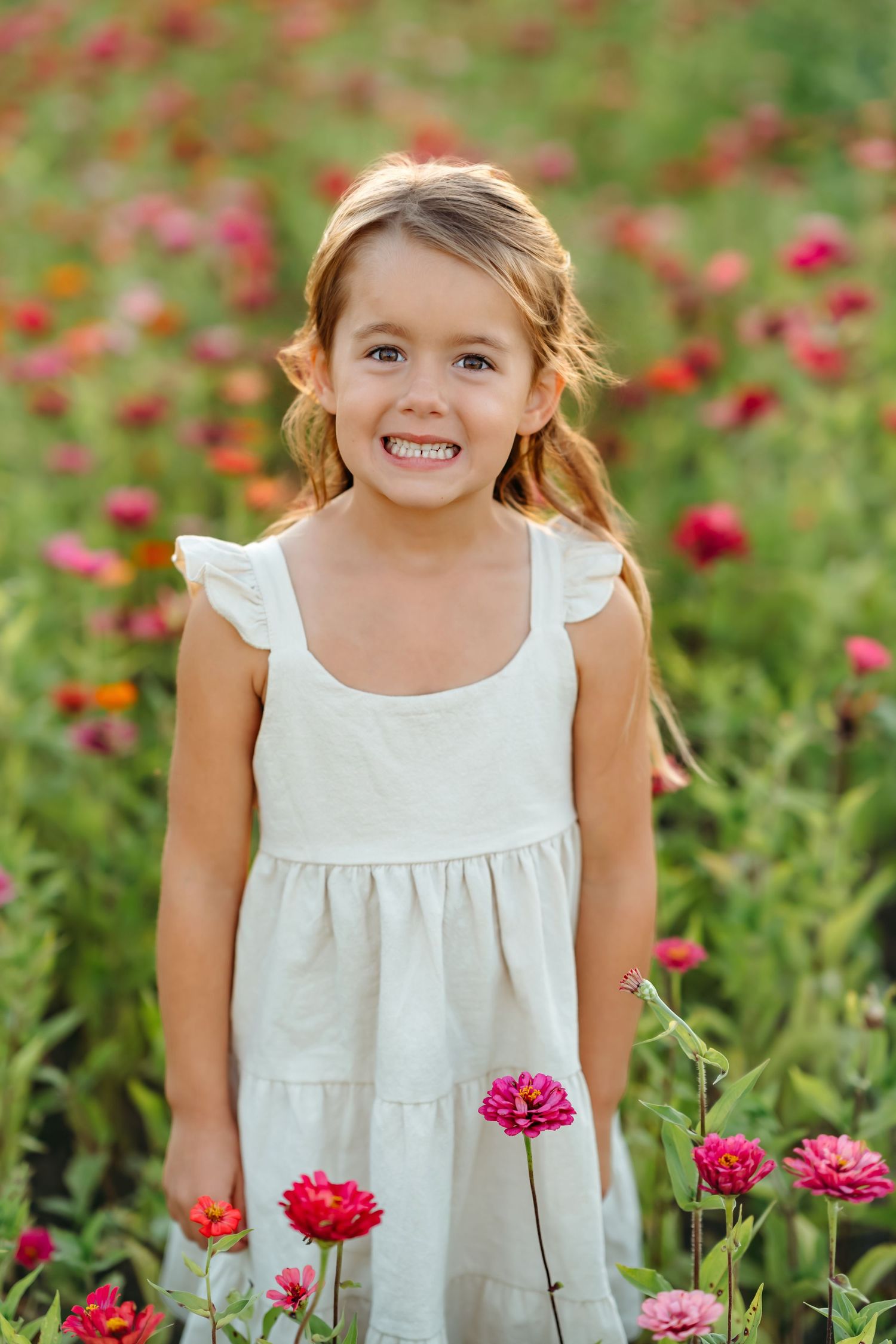 Little one in white dress smiling joyfully among bright pink zinnias in a garden setting.