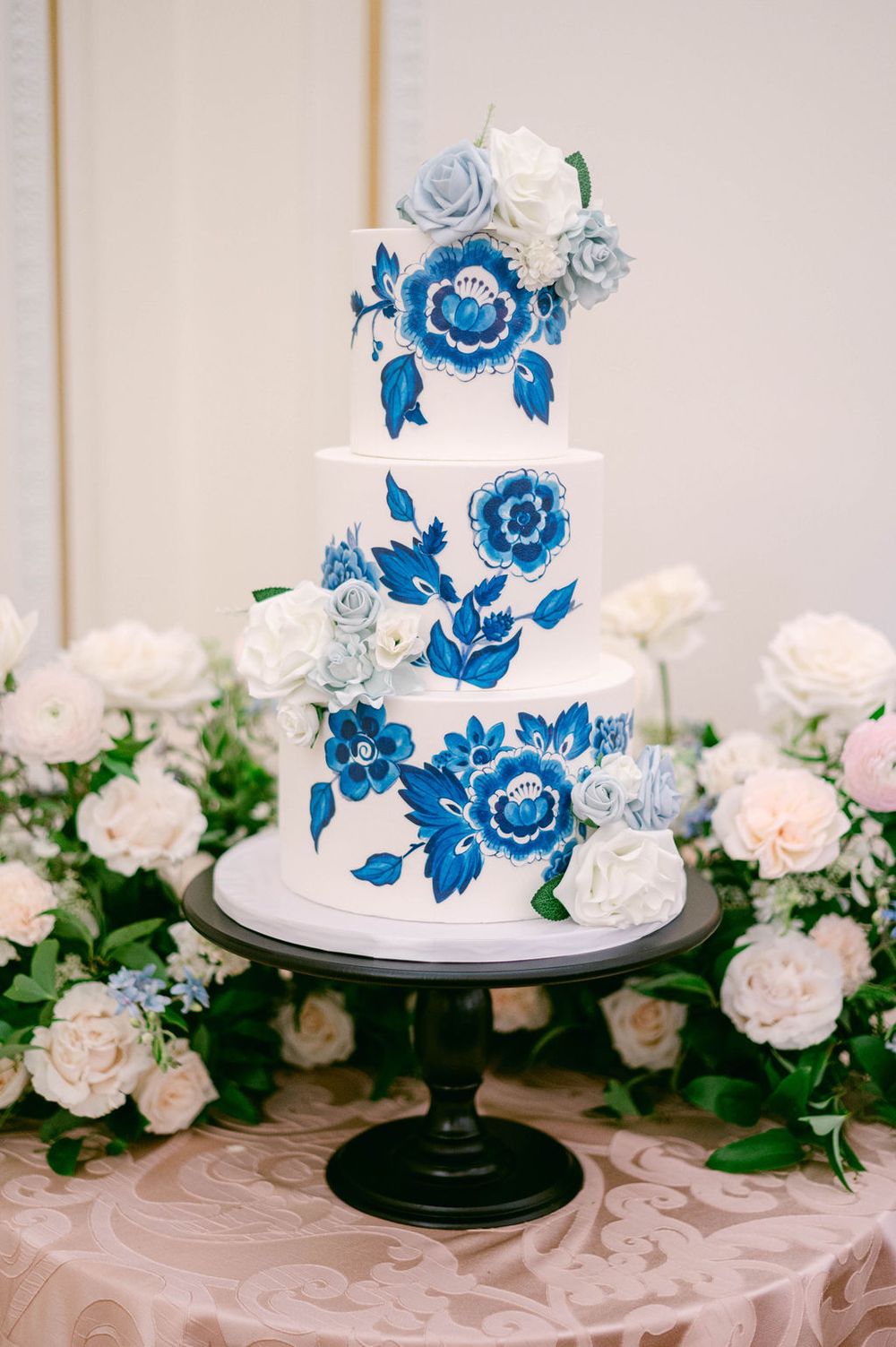 White wedding cake decorated with elegant blue floral designs and sugar flowers sits on a black stand against a floral backdrop.