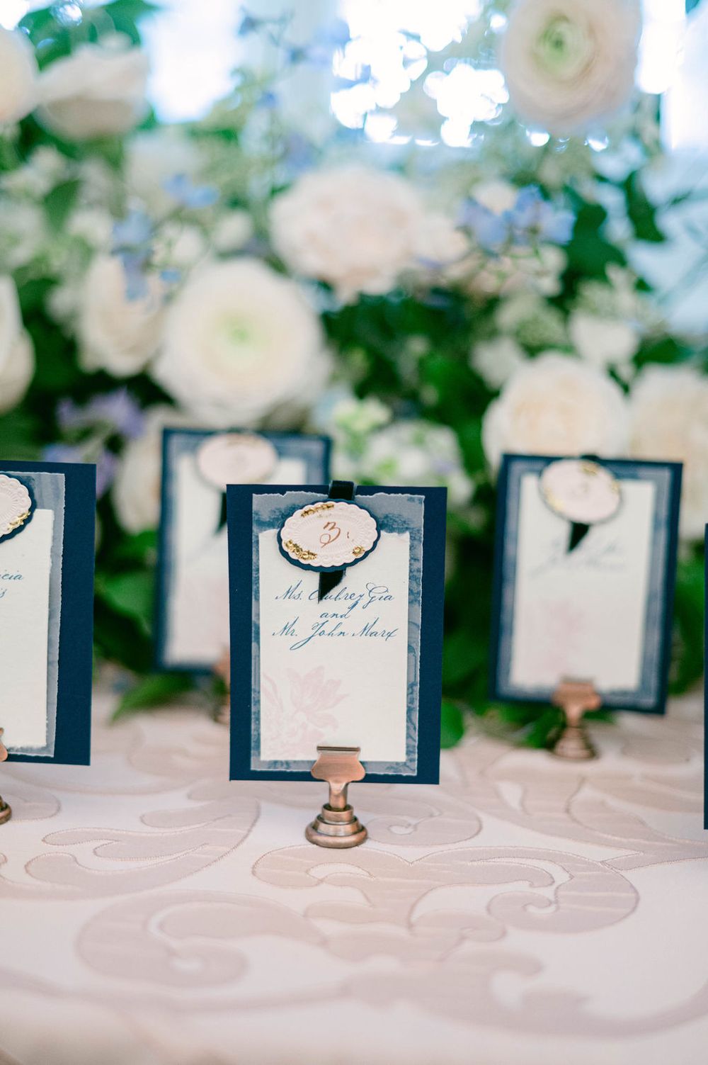 Wedding place cards arranged in navy blue holders on a textured white tablecloth with floral decorations.