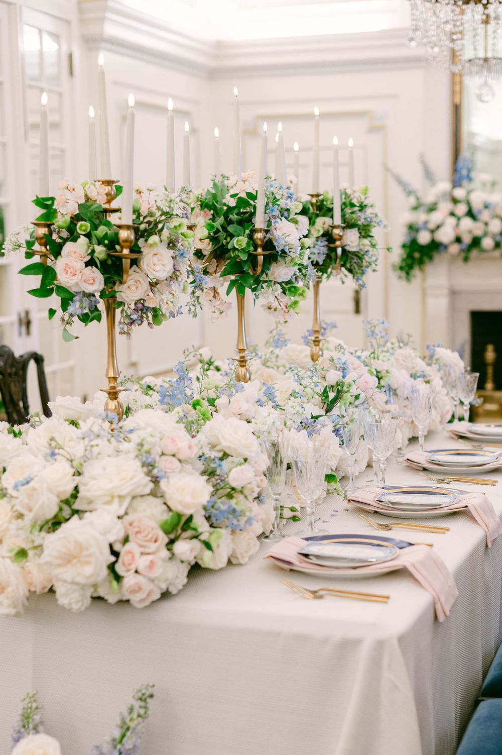 Long dining table with lush white roses, blue hydrangeas, and brass candlesticks in a bright white room.