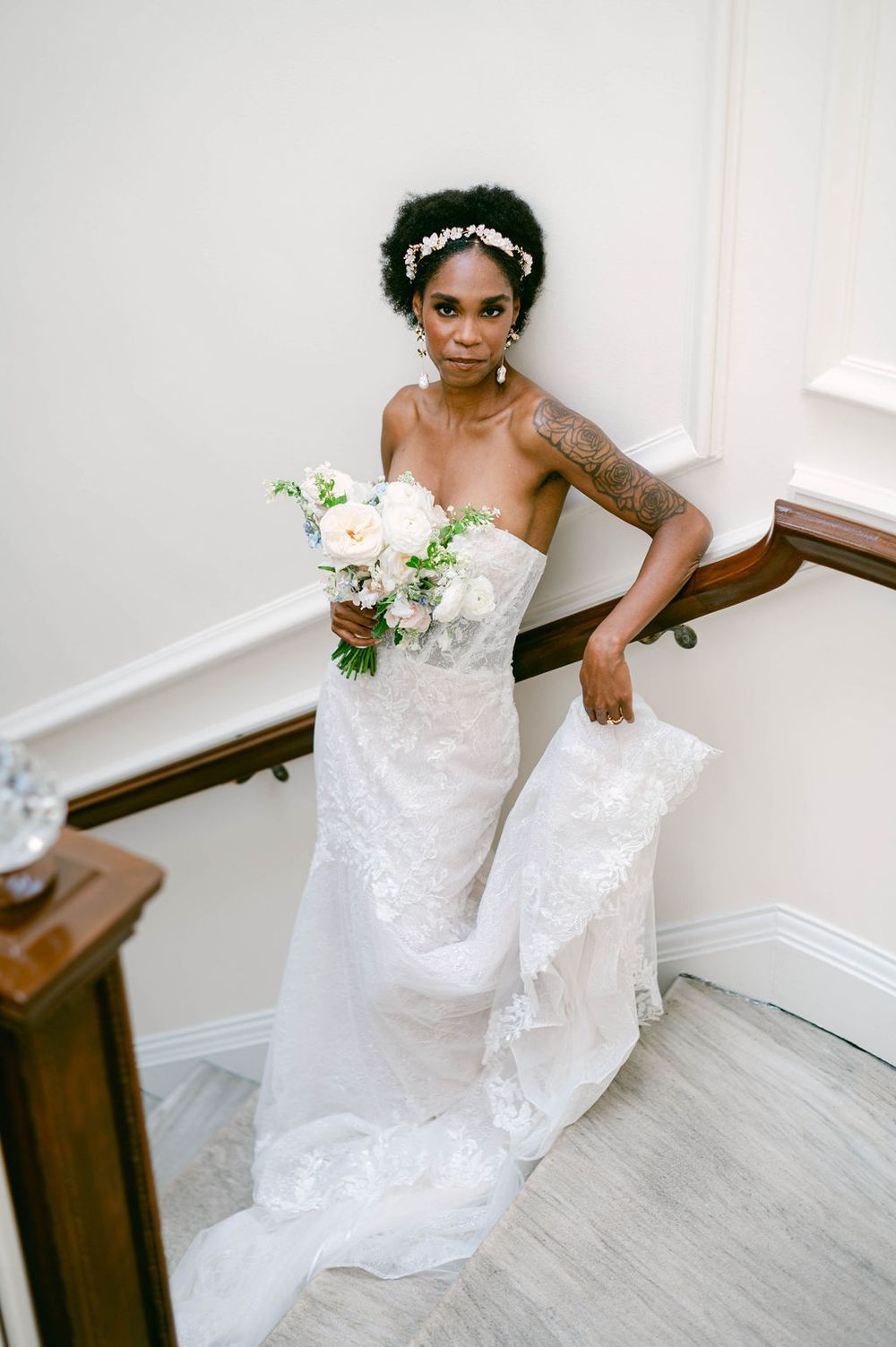 Bride in a stunning white lace gown poses gracefully against a bright interior wall with wooden banister.