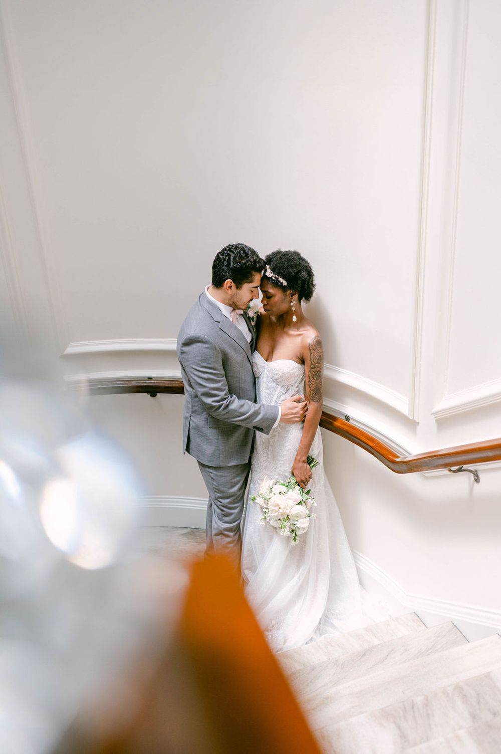 A bride and groom share an intimate moment on a staircase in a white interior setting.