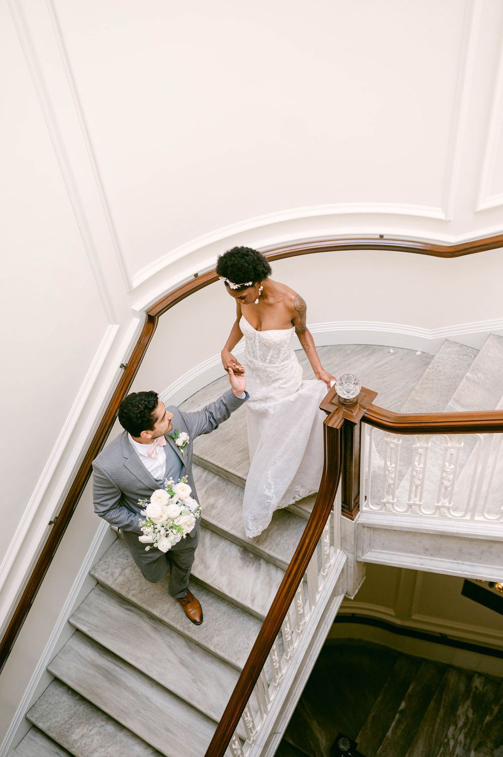 A newlywed couple descends an elegant marble staircase with white walls and wood railing in romantic poses.