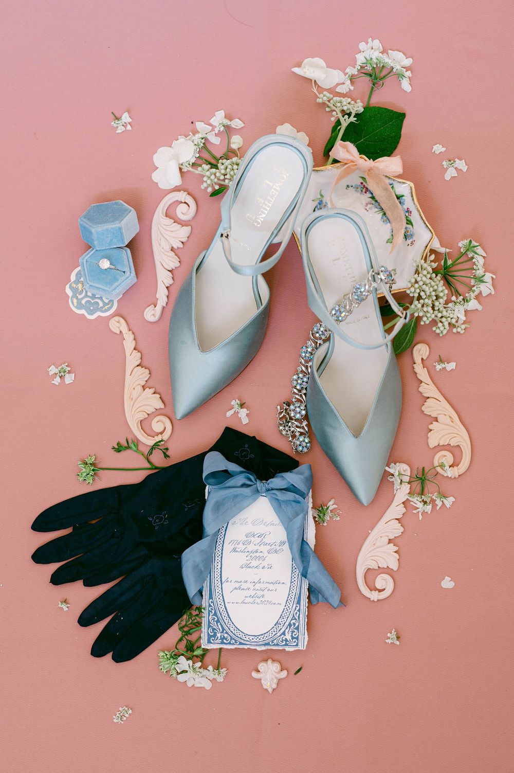 Elegant blue wedding shoes styled with black gloves, floral details, and ornate paper decorations on pink background.