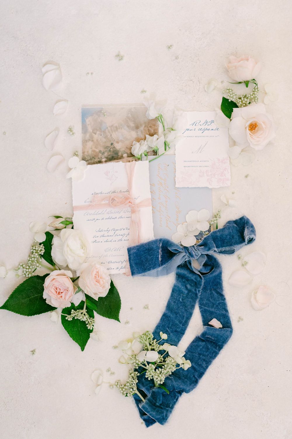 Delicate wedding stationery and blue silk ribbons styled with blush roses and greenery on a light marble surface.
