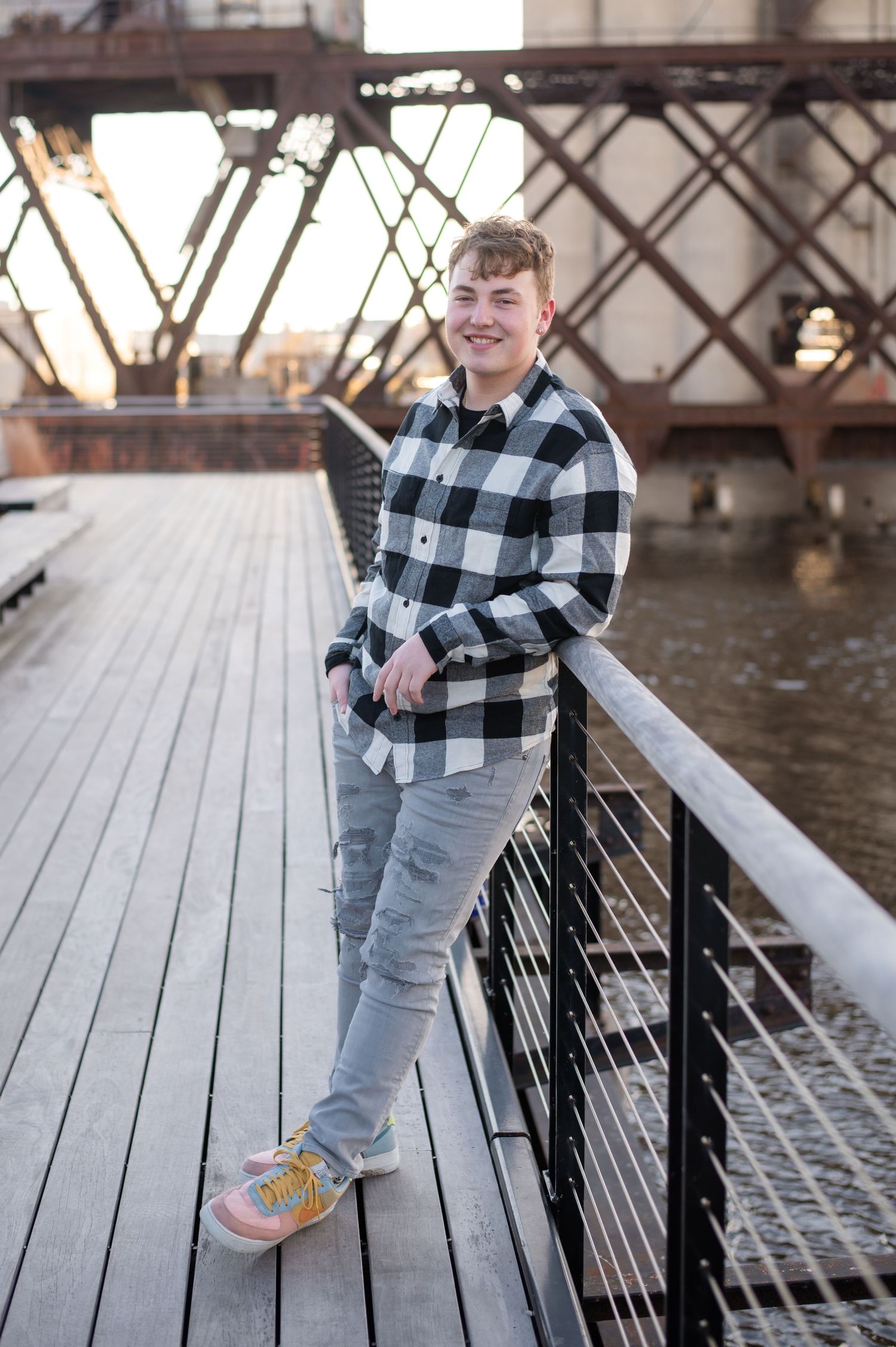 Person in buffalo plaid shirt and light jeans poses casually on wooden bridge walkway at sunset.