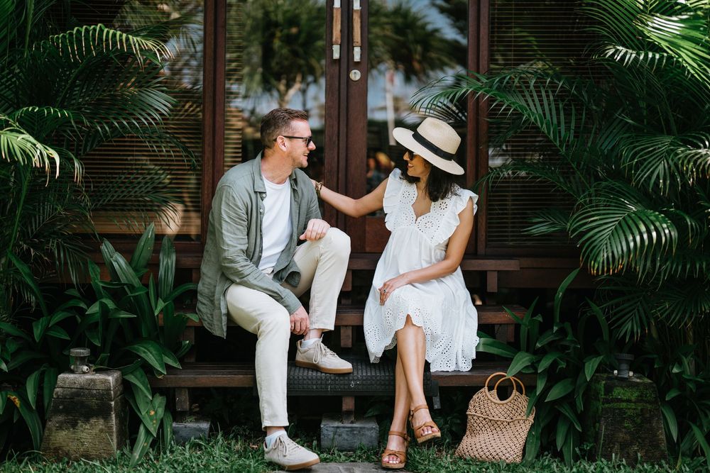 A couple sits on a wooden deck surrounded by tropical plants and palm fronds.