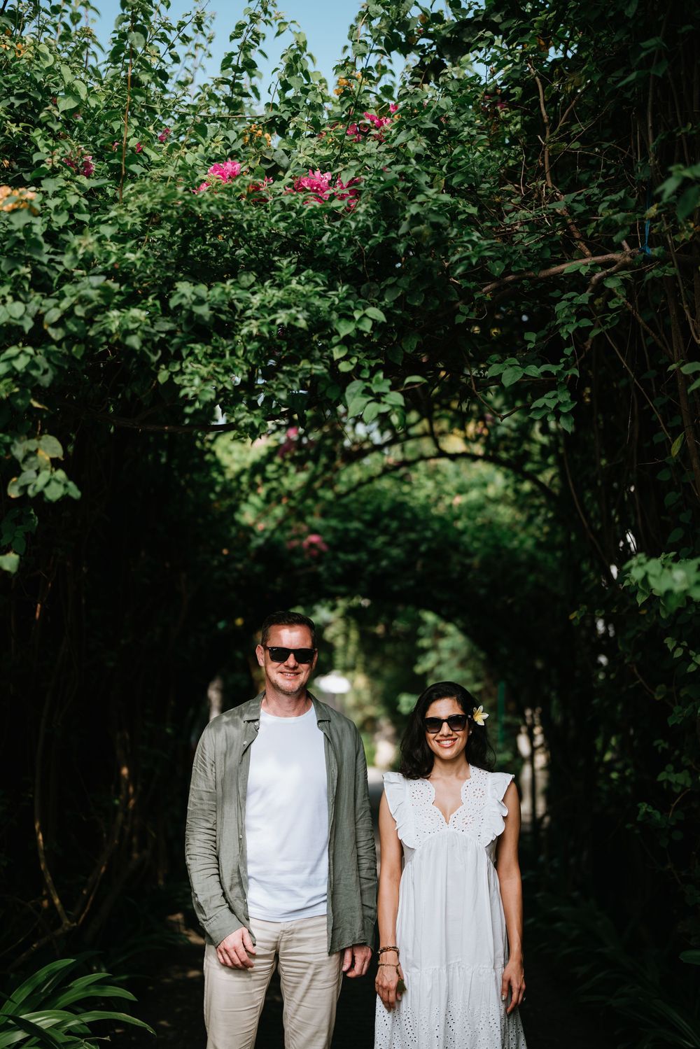 A couple stands under a floral archway with pink blooms in a garden setting.