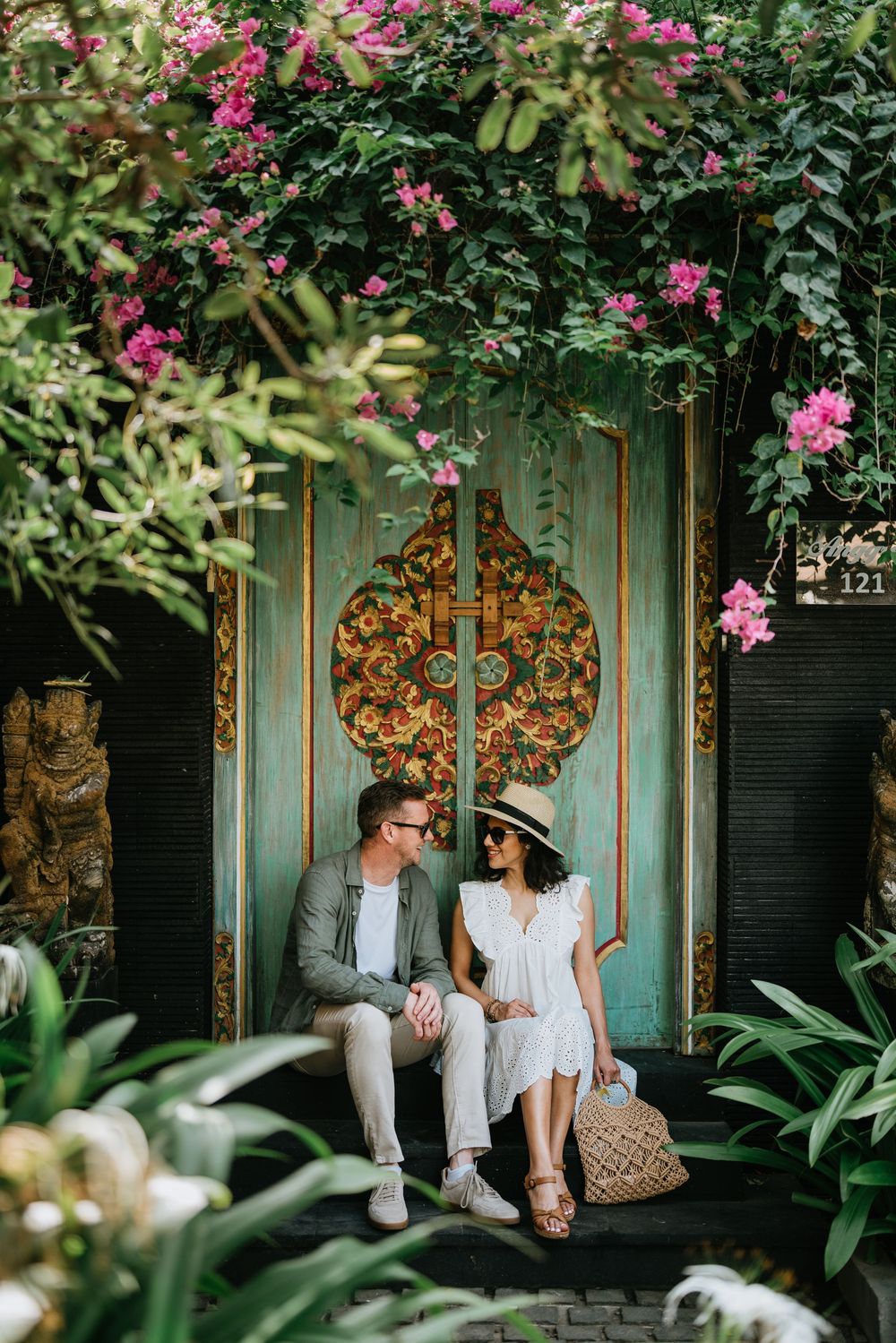 A couple sits on a bench by a decorative turquoise door surrounded by pink flowers.