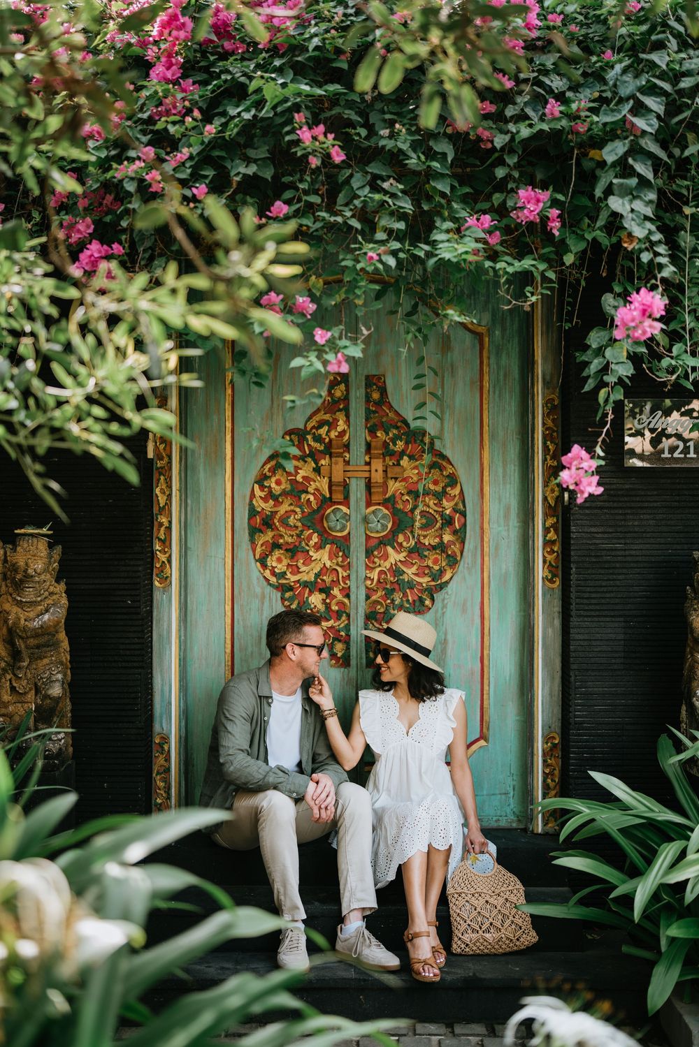 A couple sits on a bench in front of an ornate turquoise door surrounded by pink flowers and tropical foliage.