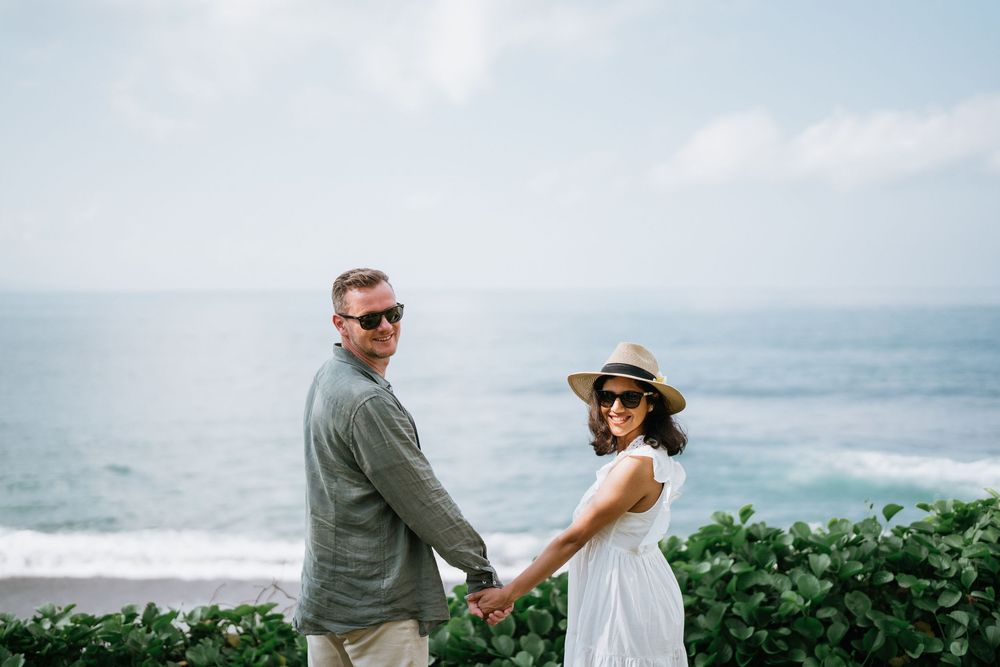A romantic couple holds hands while standing on a cliff overlooking the ocean in tropical setting.