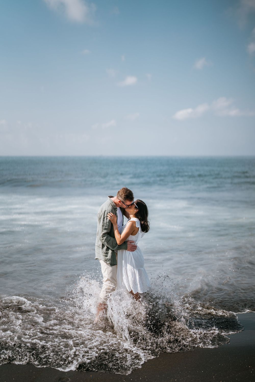 A couple shares an intimate moment as ocean waves crash around them on a black sand beach.