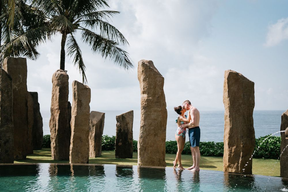 Two people share a romantic moment by a modern stone sculpture pool with palm trees and ocean views.