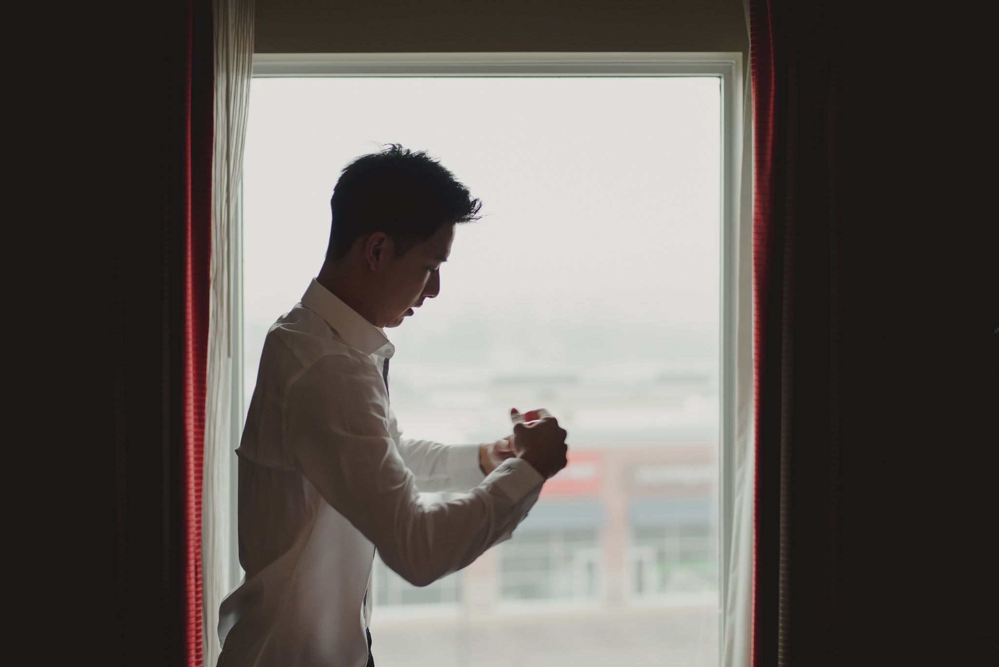 A silhouetted figure stands by a window in a white shirt, looking down at their clasped hands in contemplative pose.