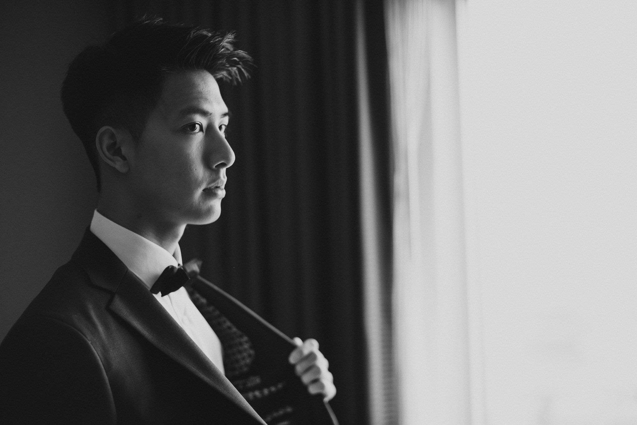 A person in a black tuxedo and bow tie stands in profile near a window in a dramatic black and white portrait.