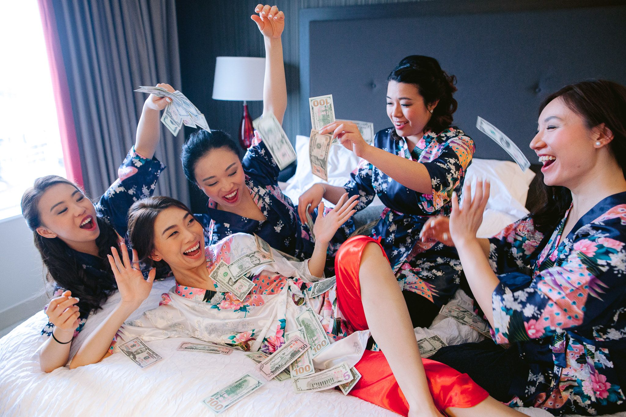 Bridesmaids in floral robes celebrate joyfully on a bed during wedding preparations.