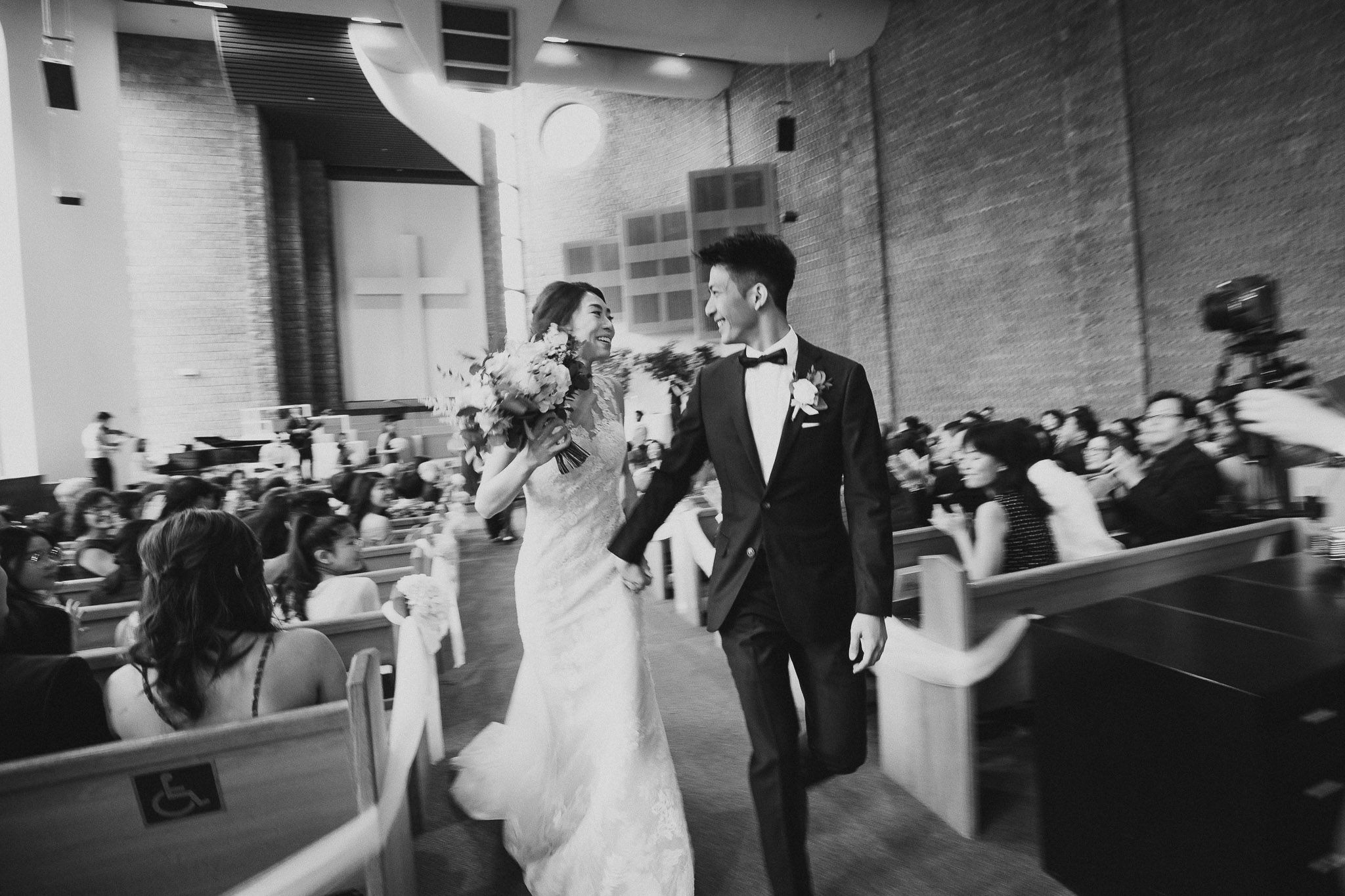 Newlyweds walk down the aisle together in a church filled with guests, captured in black and white.
