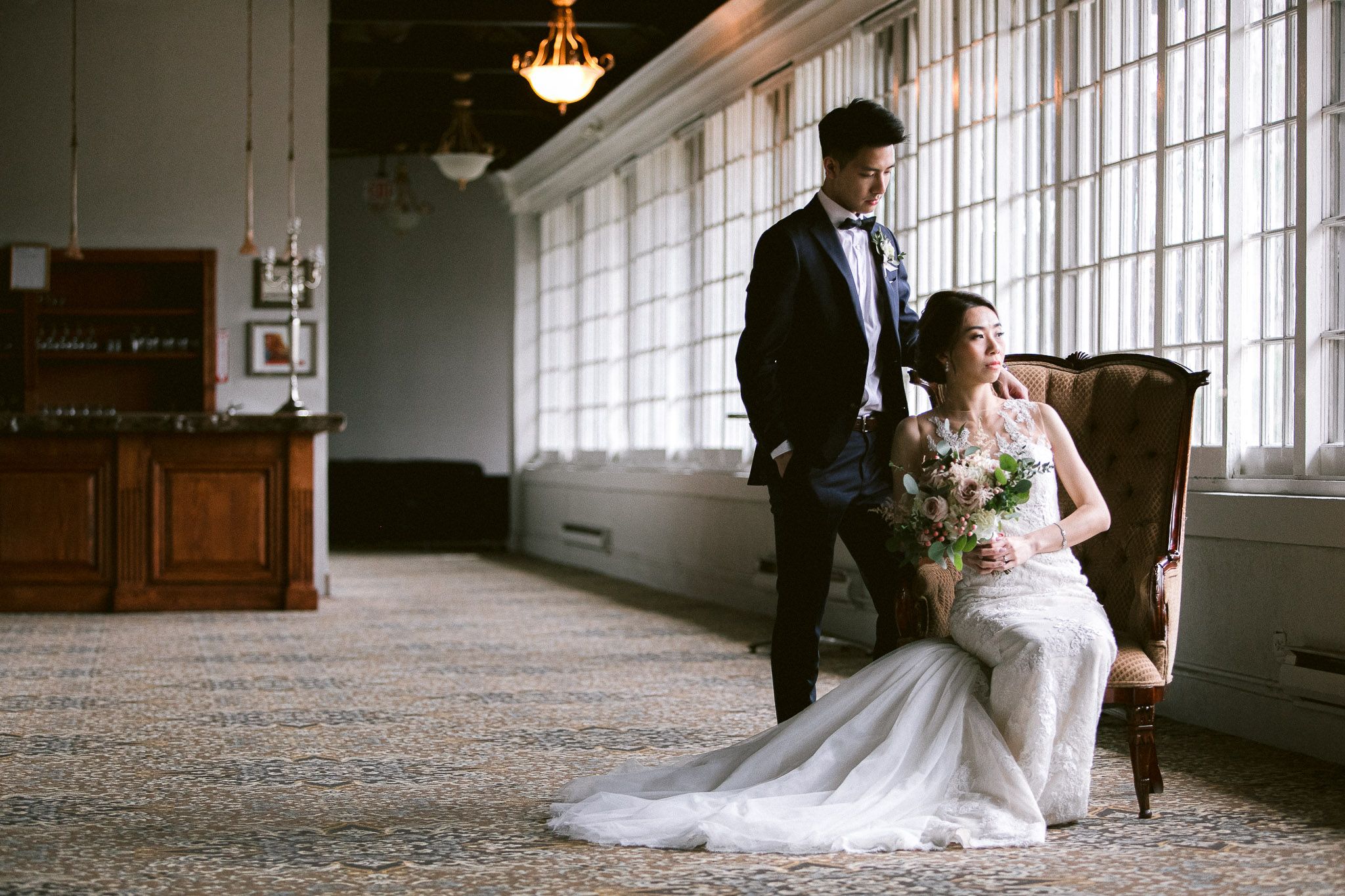 A couple poses in an elegant venue with large windows and hardwood floors.