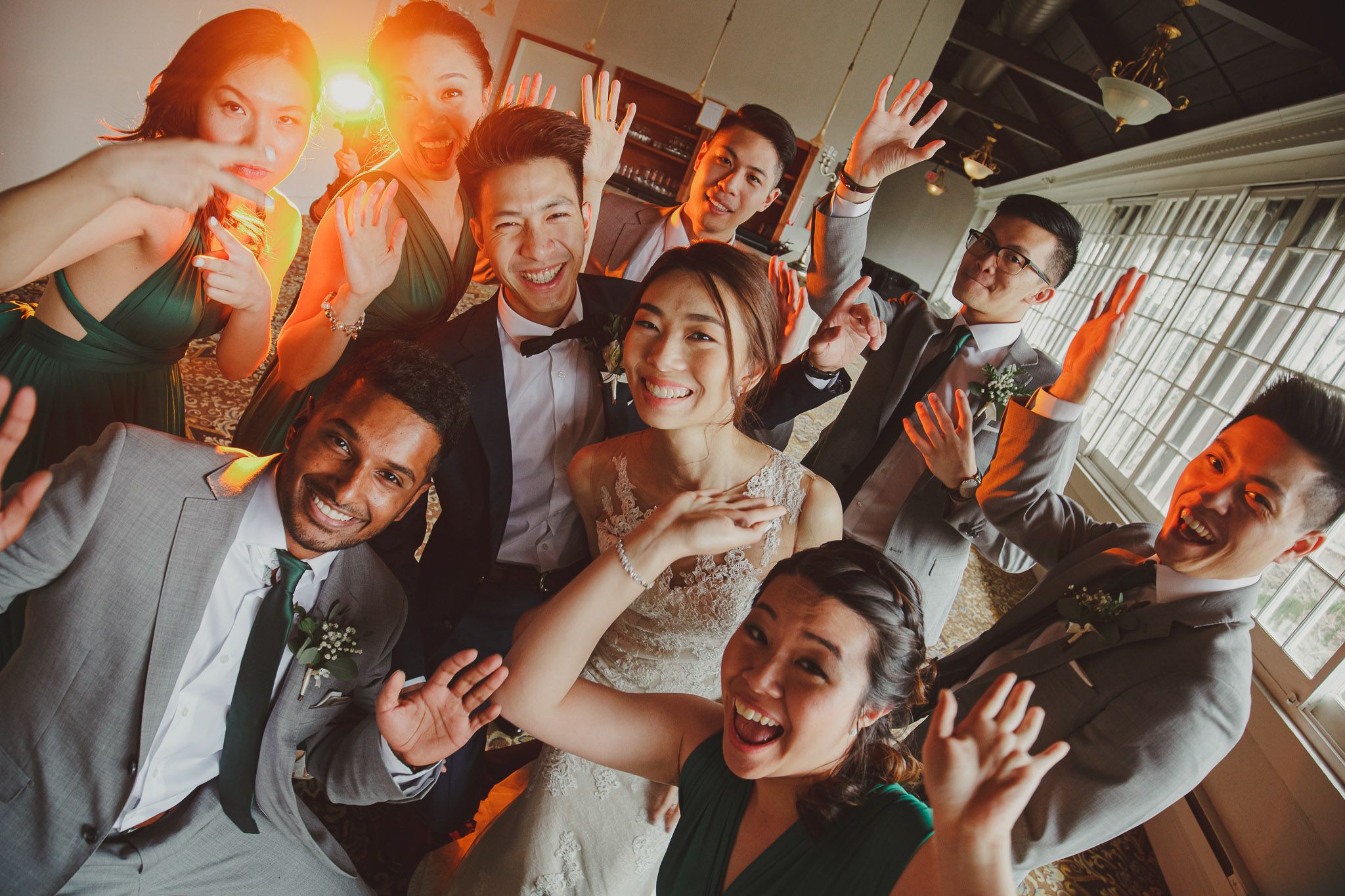 A joyful wedding party poses together in formal attire with energetic expressions and raised hands.