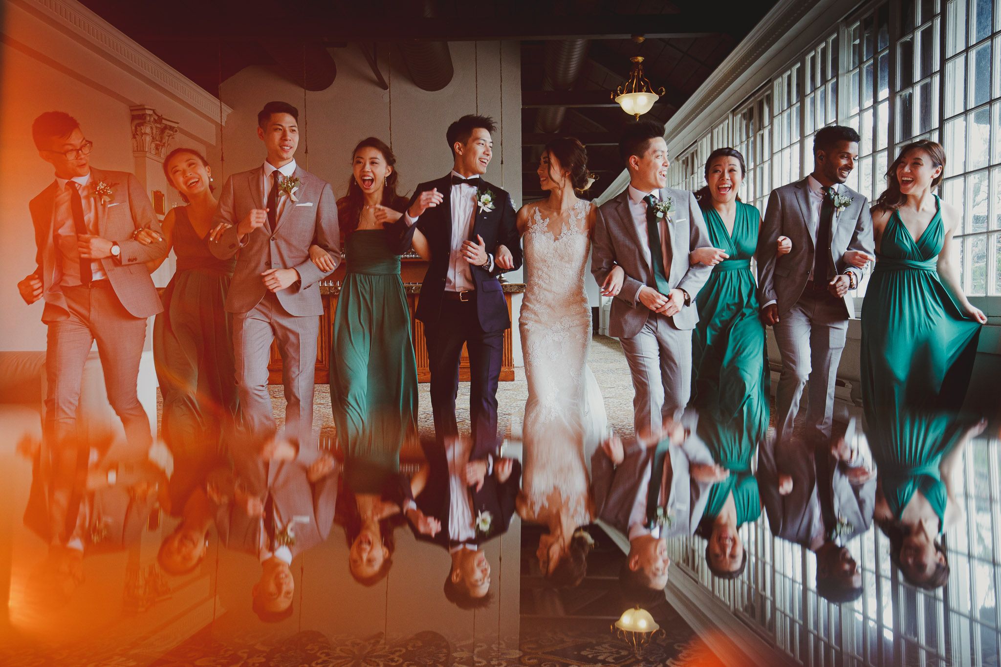 Wedding party members in teal dresses and dark suits pose in a reflective hallway.