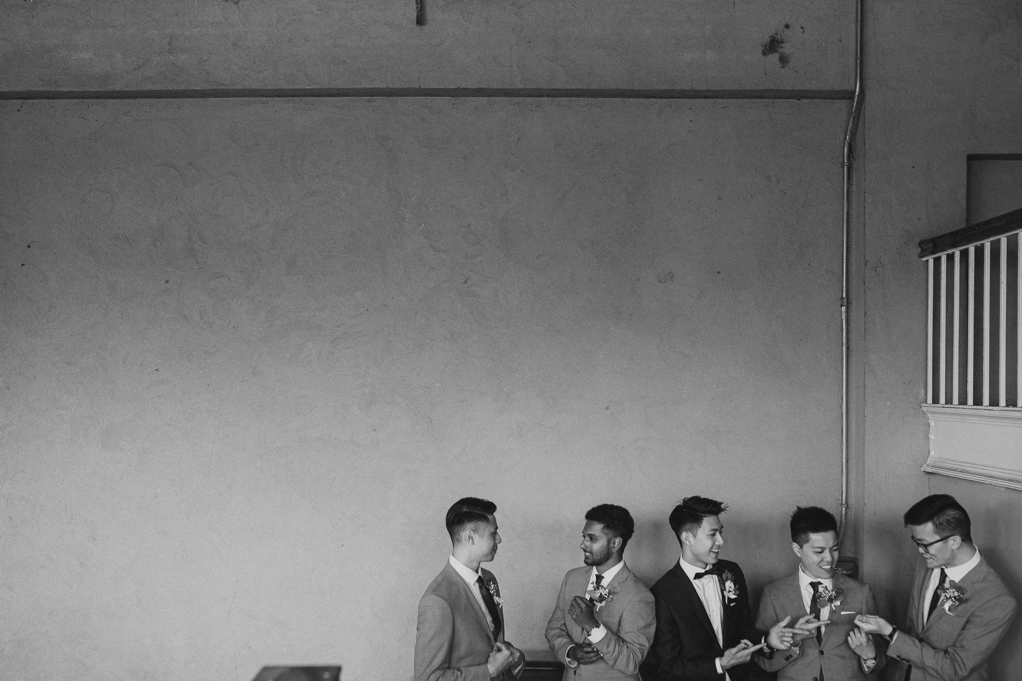 Groomsmen in formal attire stand in line against white wall at wedding venue.