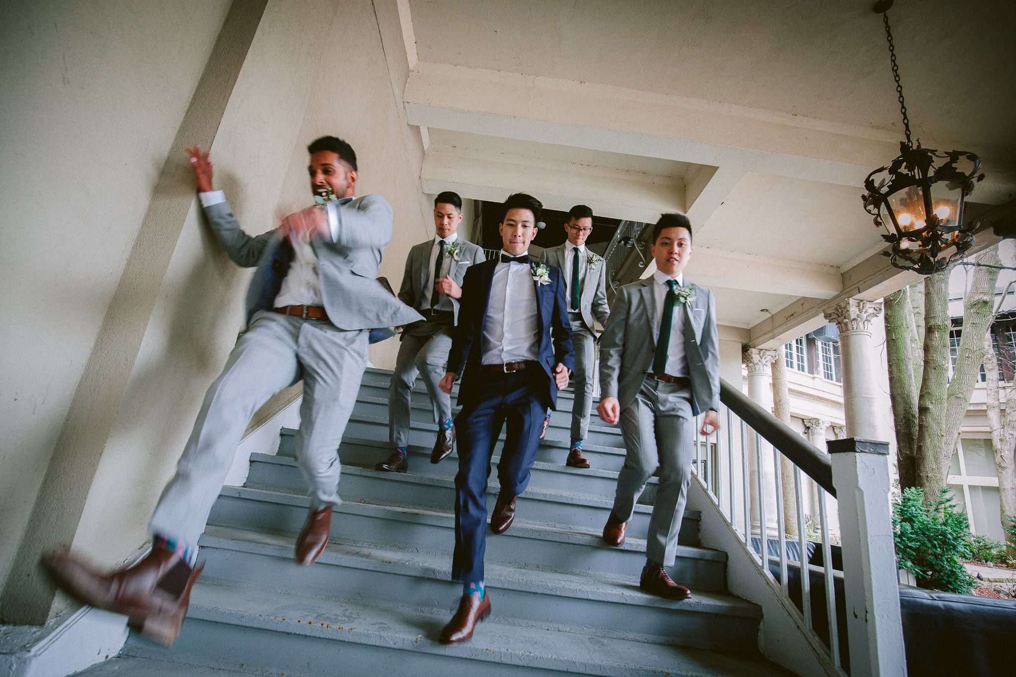 Groomsmen run and jump playfully down a bright hallway in matching suits.