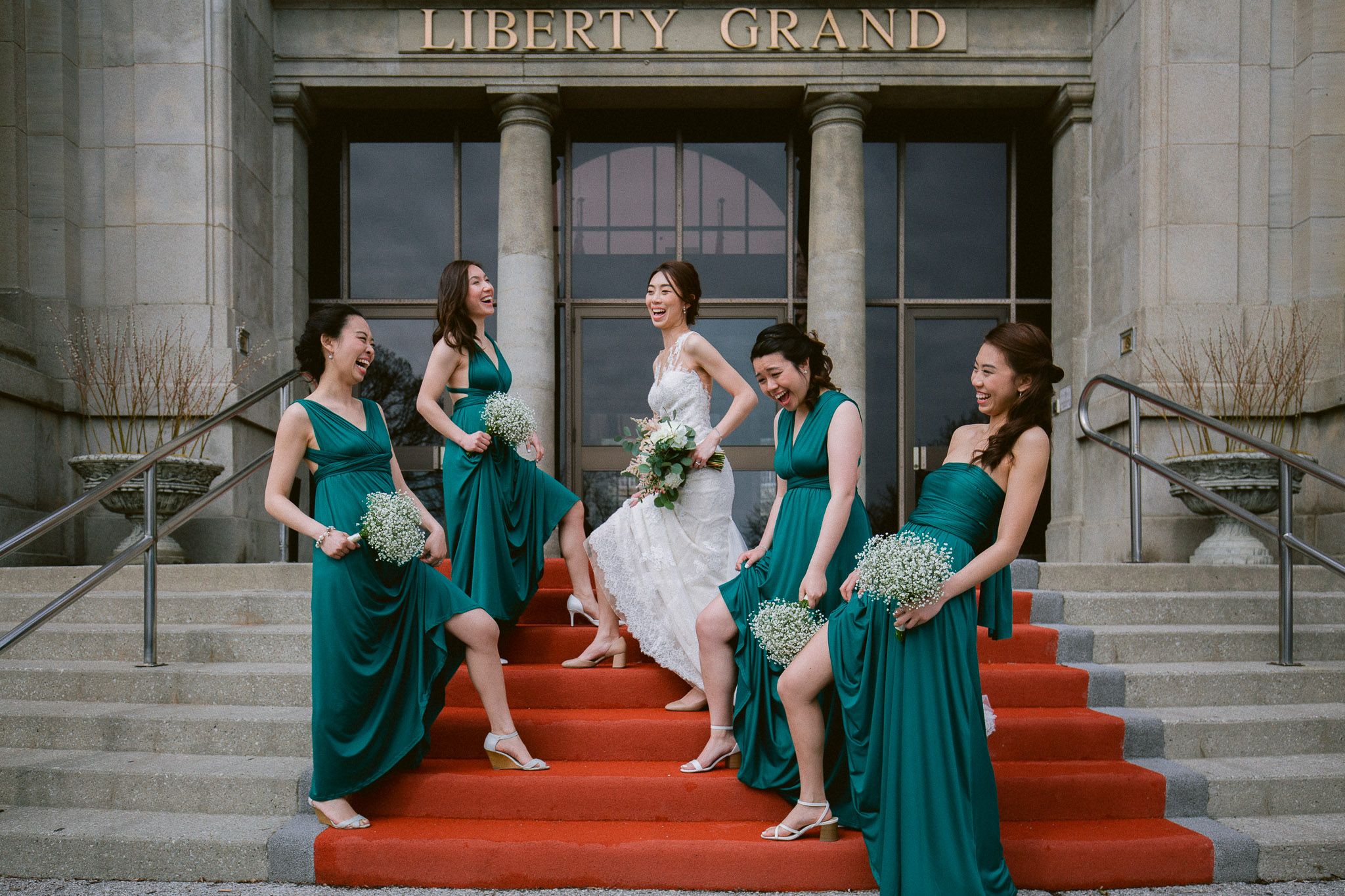 Wedding party relaxes on red carpeted steps outside the Liberty Grand building entrance.