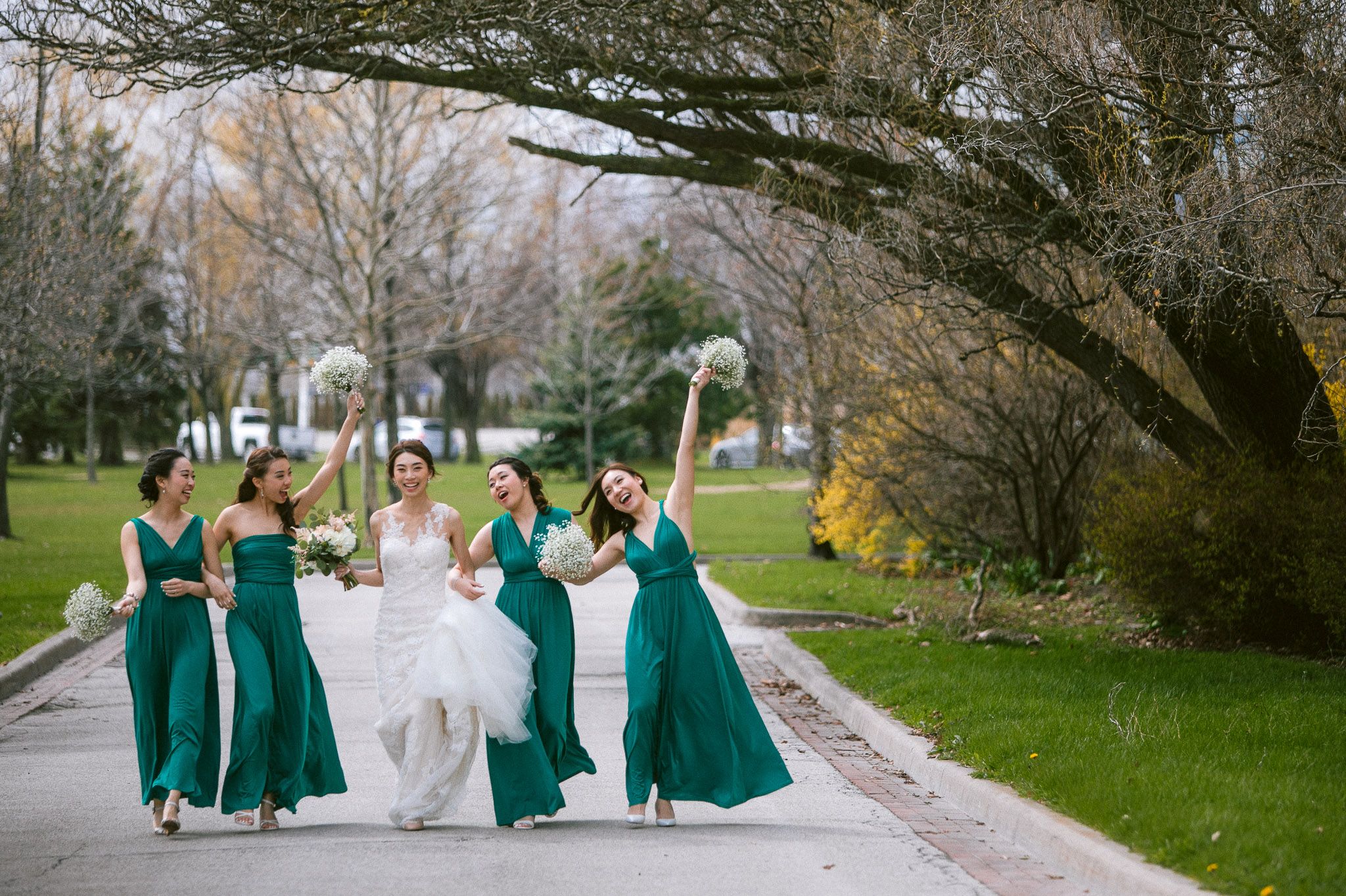 Wedding party in emerald green dresses and white gown walk together down a tree-lined path in springtime.