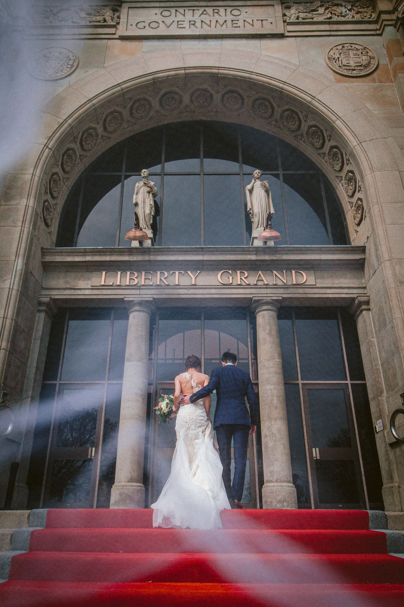 Wedding couple poses on red-carpeted steps at the historic Liberty Grand Ontario Government building.