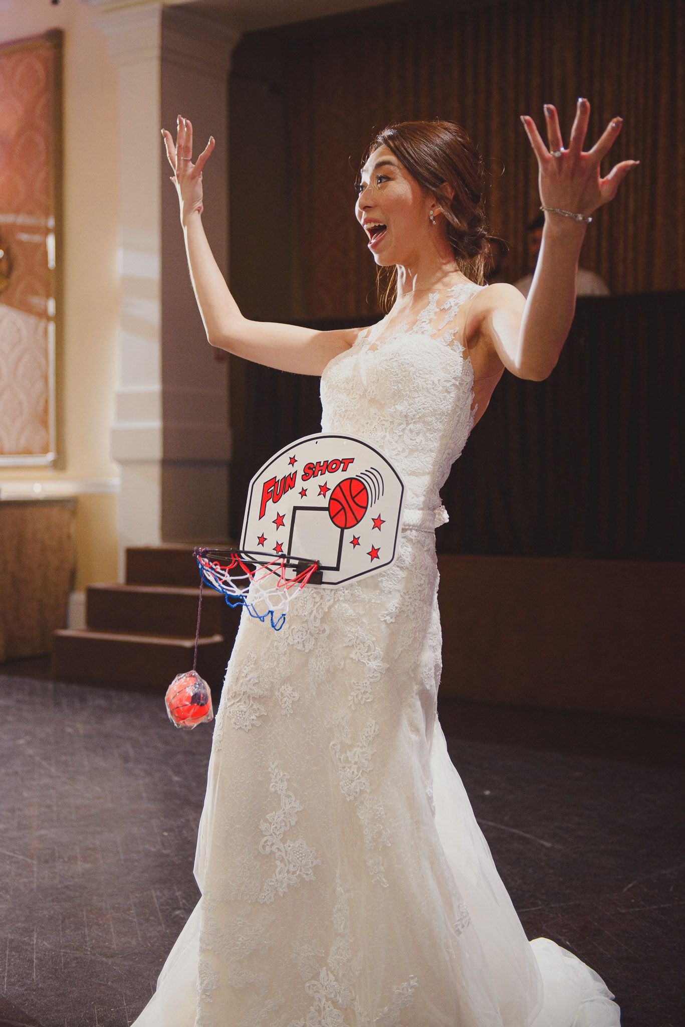 A bride in a lace wedding dress poses playfully with a basketball hoop prop during her reception.