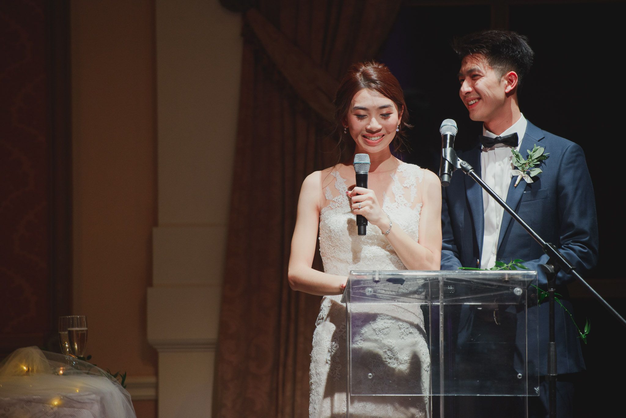 A couple speaks at a wedding reception podium with elegant decor and lighting.