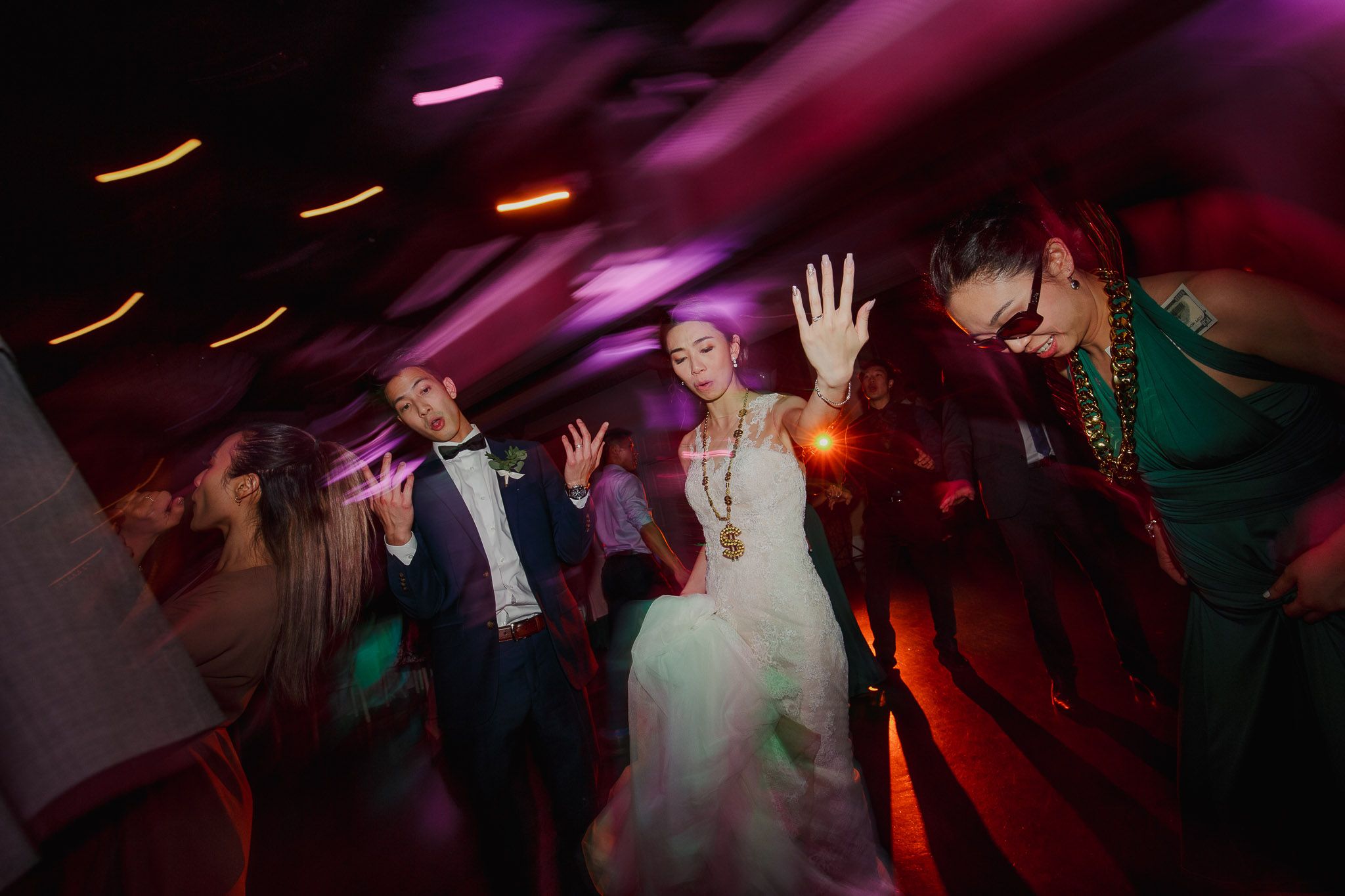 Wedding guests dance and celebrate on a dimly lit dance floor with purple and red lighting effects.