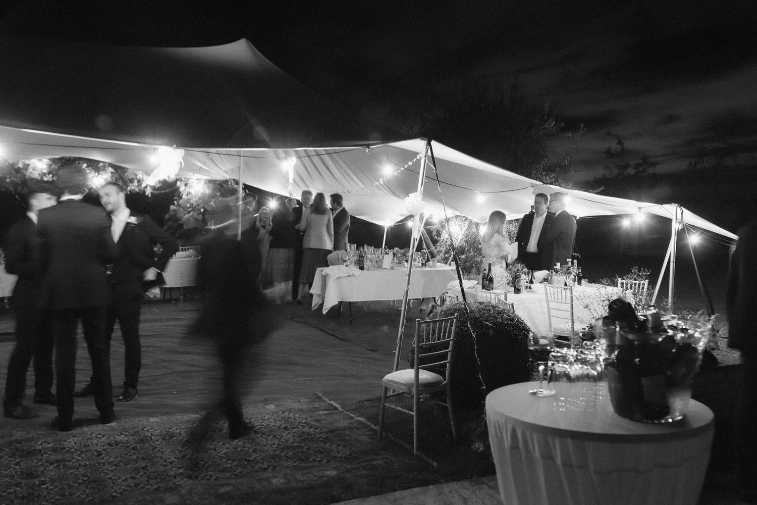 Black and white photo of an outdoor evening reception under a lit tent with guests mingling and tables set for dining.