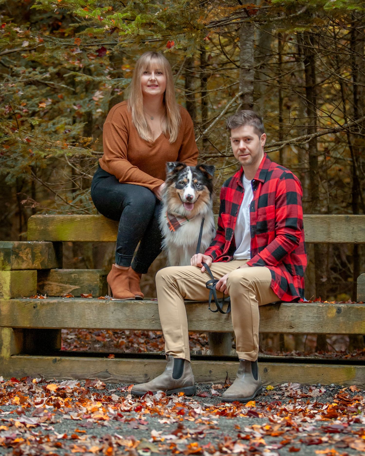 People sit on a wooden bench with their Australian Shepherd dog surrounded by colorful autumn leaves in a forest.
