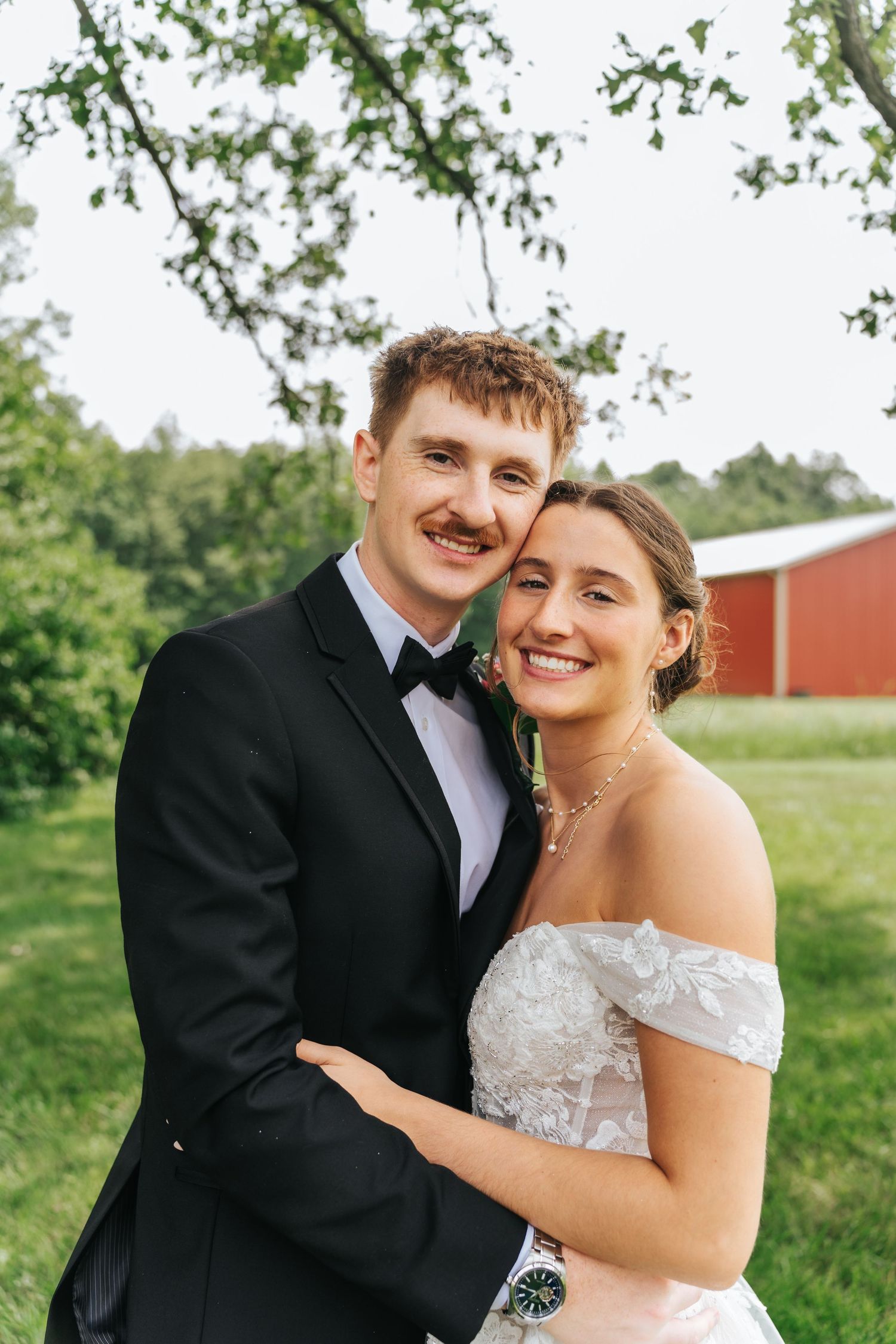 A couple in formal wedding attire smile together outdoors with a red barn and green field in the background.