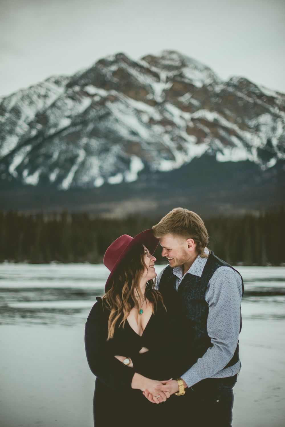 A romantic couple embraces against a stunning backdrop of snow-capped mountains and a frozen lake. Jasper Engagement Photographer