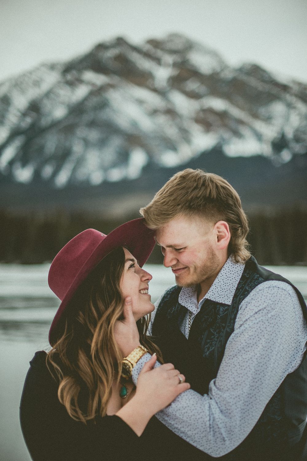 Romantic winter engagement session with snowy mountain backdrop as couple shares intimate moments at dusk. Jasper Engagement Photographer