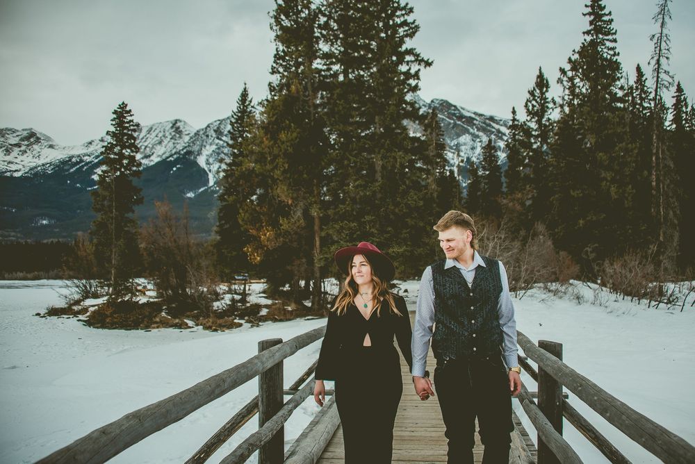 Couple walking hand in hand on a wooden boardwalk surrounded by snowy mountains and evergreen trees Jasper Engagement Photographer