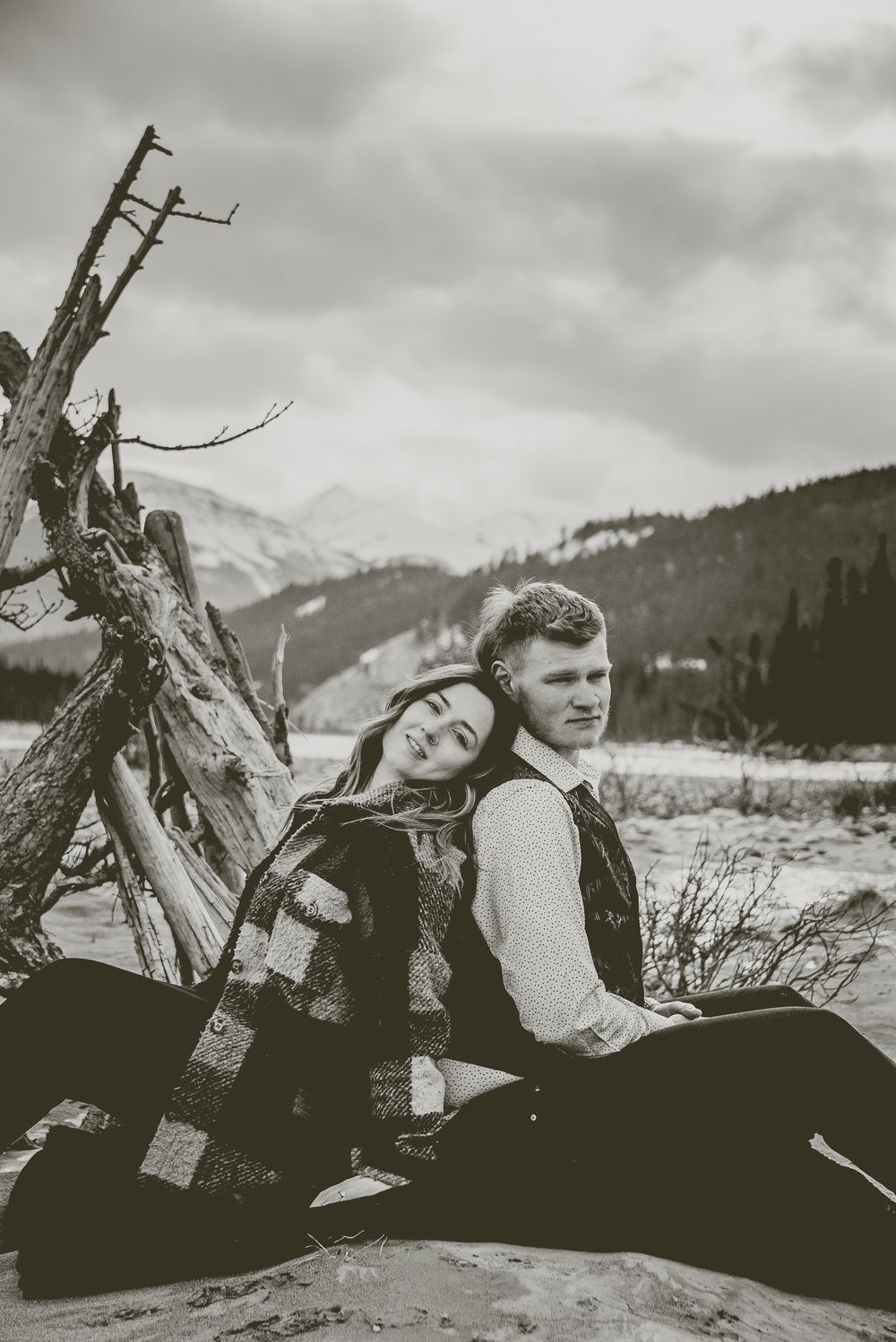 Black and white portraits show a couple lying in a meadow with mountain backdrop and dead tree Jasper Engagement Photographer