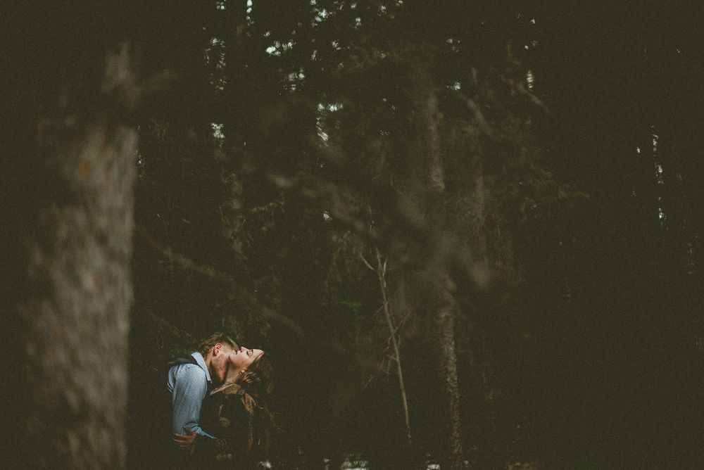 Two people share an intimate moment while standing in a dark wooded forest setting. Jasper Engagement Photographer
