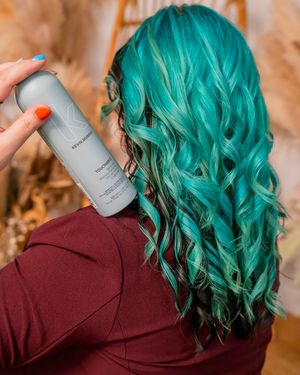 Person with vibrant teal hair holding styling product against burgundy top.