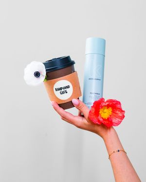Hand holding a coffee cup and red flower against white background.
