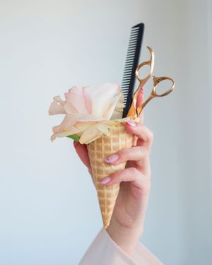 Hand holding a comb and scissors with an ice cream cone prop against white background.