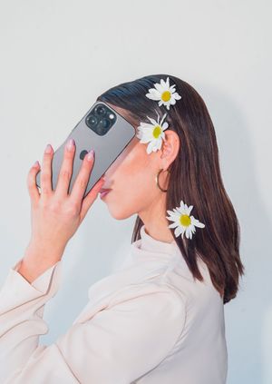 Person wearing white with daisies in hair holding phone against light background.