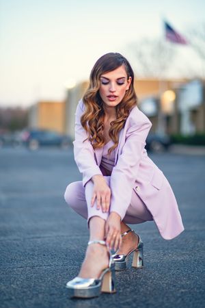Fashion model in pink suit and silver heels crouching on asphalt.