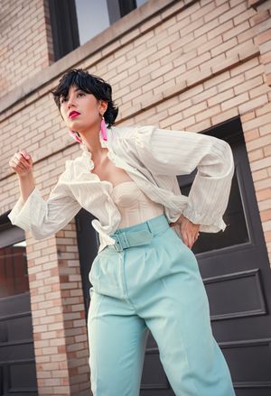 Model wearing ruffled white blouse and mint pants poses expressively against brick wall.