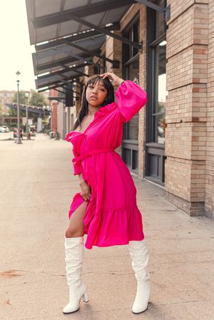 Model in hot pink dress and white boots poses confidently outside brick building.