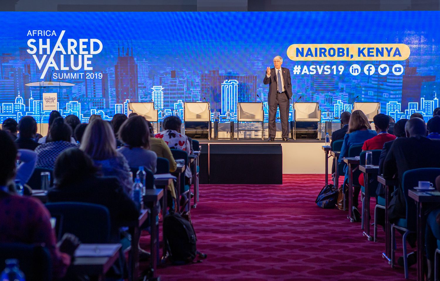Conference attendees seated in rows of chairs with red upholstery face a brightly lit stage during a business event.